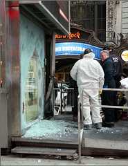 Officers and members of a bomb squad unit inspect the damage. The explosion left a gaping hole in the front window and shattered a glass door, twisting and blackening the metal frame of the building.