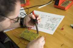 A close-up of a person holding a black and red tool that's touching a green object covered in silver wires. A detailed drawing is also on the table.
