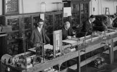 Black and white photo of men in suits standing behind a large, wooden table with lots of wires and spools.