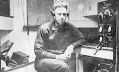 Black and white photo of man seated and is surrounded by radio equipment.