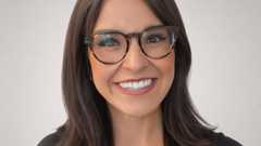 A woman with long dark hair and glasses smiles warmly against a plain light gray background.