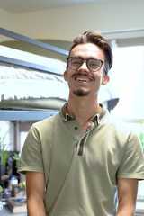 A young man with glasses and a goatee smiles while standing in a room with a bunk bed and a window behind him.