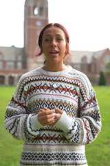 A smiling woman with red hair stands on a green lawn wearing a patterned sweater with a clock tower and brick building.