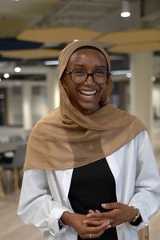A smiling person wearing glasses, a tan headscarf, a white shirt, and a black top stands indoors with hands clasped.