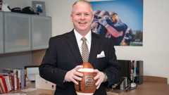 A man in a dark pinstripe suit smiles while holding a football in an office with sports photos and books.