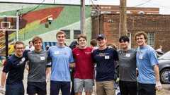 Seven young men stand arm in arm smiling outdoors near a basketball hoop and a colorful mural on a brick wall.