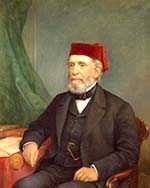 White man with grey beard, wearing suit and red fez, seated in a wooden armchair and resting his elbow near books on a table.