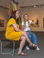 Two students sitting on a bench in an art gallery looking at framed artwork with one student pointing.