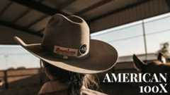 A man wearing an American Hat Company 100x felt hat is seen from behind looking off at a dirt ground with overcast skies.