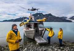 Crew members of a scientific expedition send a drone near their vessel, docked at an icy, rocky landscape