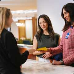 Two Mayo Clinic staff members greeting a patient in a bright lobby area, smiling and exchanging paperwork.