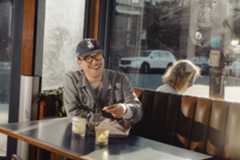 A man wearing glasses and a cap smiles while eating at a table with a drink and candle, sunlight streaming through large windows behind him.