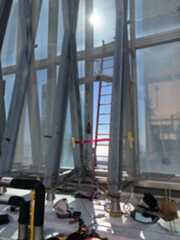 The image shows a construction site with metal beams, a red ladder, and various tools and materials scattered on the ground. Sunlight shines brightly through a mesh screen.