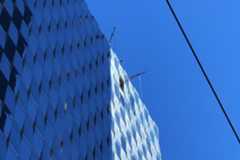 A glass-covered high-rise building reflects the blue sky, while two window cleaning platforms are visible near the top, with a cable diagonally across the scene.