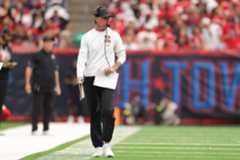 A football coach in a white 49ers shirt, black pants, and headset walks on the sideline holding papers during a game with blurred crowd in the background.