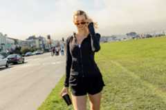 A woman in black athletic wear holds a black tumbler, adjusting her sunglasses, standing on grass beside a street with cars and people.