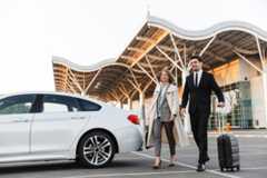 A man in a suit and a woman in a coat walk together in an airport parking lot, pulling a suitcase, with a white car parked and a modern terminal building in the background.