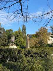 View of bronze statue of Queen Isabella from the side, from a distance. She is framed by green and yellow shrubbery and branches hang overhead, backed by a blue sky with light, puffy clouds.