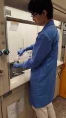 A teenager wearing a long, teal lab coat and safety glasses pouring a solution into a glass funnel.