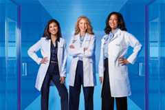 Three female doctors stand confidently in a bright blue hospital corridor. They wear white lab coats over professional attire, with two of them having their hands on their hips and the middle one crossing her arms. The background features glass doors and a tiled ceiling.