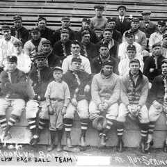 Group of white men in baseball uniforms with caps and jackets sitting on wooden bleachers