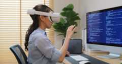 A woman sitting at a desk behind a computer works on coding project while wearing VR goggles.
