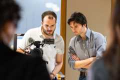 Student Media Technology Coordinator Austin Orr is on the right, wearing a blue and white striped shirt and with his arms crossed, and is leaning in to look at a the display view on a video camera that a student is holding. The student is to the left of Austin and is wearing a white shirt and is also looking at the display.