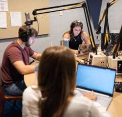 Three students sit in a WUOT studio, all looking at their laptops and two wearing headphones.