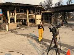 A photo of School of Journalism and Media alumna Ashley Sharp ('18) reporting on the 2025 LA Wildfires while standing in front of a burned down building.