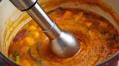 A close-up shot of a hand-held immersion blender actively pureeing vegetables in a pot, emphasizing the ease of the process.