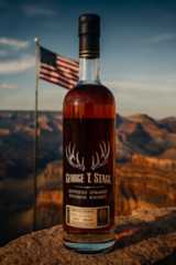 alt="ジョージ・T・スタッグ(George T. Stagg)ケンタッキー・ストレート・バーボンウイスキーのボトル。グランドキャニオンの夕景を背景に、アメリカ国旗がはためく中、琥珀色のウイスキーが輝く様子をクローズアップで撮影した写真。"