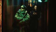 Pop star Charli XCX wears thick black sunglasses and a puffer jacket as she walks with her arms crossed down a dark, green-lighted hallway. A young woman follows closely behind with a tablet in hand.