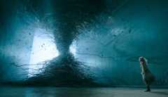 A young woman with long bleached hair and an off-white flowy dress stands in front of what seems to be a tornado of tangled wires.