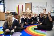 Female teacher holding up a book in front of her class of elementary school kids sitting on the floor in a classroom, side view