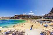 Beautiful sunny day on Amadores Beach with tourists relaxing on loungers and swimming in clear Atlantic Ocean on background of rocky hill.