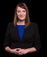 Helen farrell triangle foreground min Helen farrell in black blazer and blue top, hands clasped.