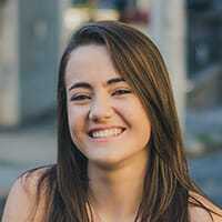 Young woman with long brown hair smiling outdoors in a city setting.