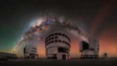 A glowing arch of the Milky Way galaxy can be seen in a red and orange and green night sky over three large towering buildings. 