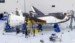 A large black and white space shuttle gets worked on by crews inside a clean warehouse