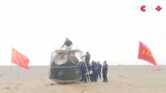 A team of people examine a metal spacecraft in the middle of a sandy area with Chinese flags waving nearby