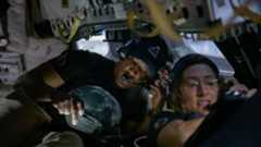 A man and a woman hunch over in a small capsule, both looking at separate electronic screens