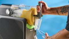 the inoviva slushie machine, a slushie maker with a plastic cylinder body, a dispensing spout, photographed making orange juice slush against a blue tom's guide background