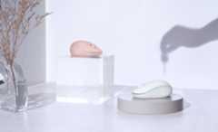 Two contoured wireless computer mice, one in dusty pink and one in mint green, are displayed on a clear acrylic block and a round grey pedestal in a brightly lit, stylized setting.