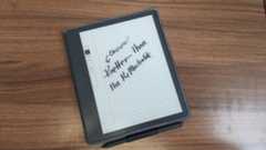 The Kindle Scribe on a woodgrain desk, with writing on the display that says &quot;cheaper than the reMarkable&quot;