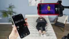 An image showing behind the scenes of our temperature regulation tests on the Saatva Classic mattress, with our tester lying on the mattress and our Managing Editor of Sleep, Claire Davies&#039; iPhone attached to our thermal imaging camera to track the heat absorption and heat dissipation capabilities of the Saatva Classic over a period of 30 minutes.