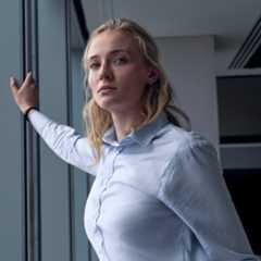 sophie turner as zara wearing a light blue button down leaning against a window in an office building in a still from steal