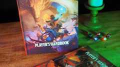 Dungeons &amp; Dragons Player's Handbook standing on a wooden table beside dice, a candle, and the 2014 Player's Handbook