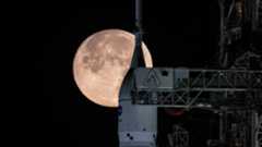 The tip of NASA's Space Launch System rocket is visible with the full moon rising behind it in the black night sky. The steel framework of the launch tower dominates the right of the screen, and the crew arm is extended, bridging the gap to the rocket's white upper stage.