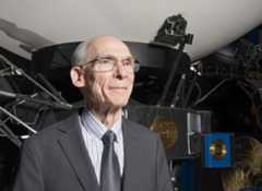 a smiling older man in a suit stands in front of a model of a large spacecraft.
