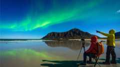 Two people sat on a beach with camera and tripod observing the northern lights.